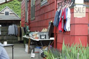 Outdoor yard sale of clothes hanging off the house and fence and many other items on tables in the driveway that has a carport over it.